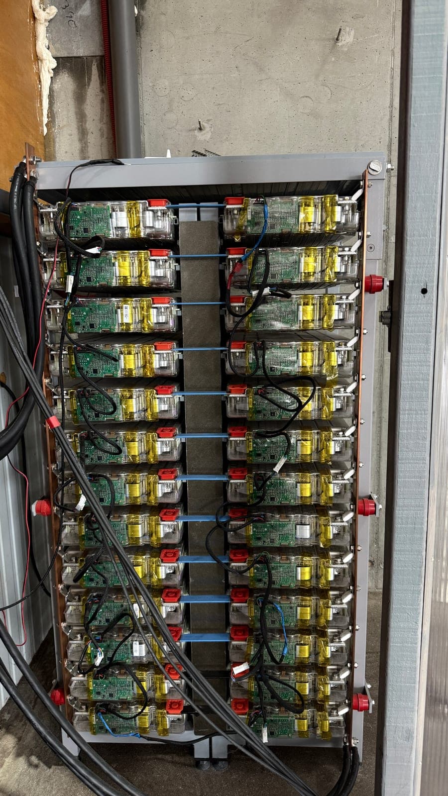 Rack de baterias com duas colunas de módulos empilhados, cada um com uma placa eletrónica verde à vista, e barramentos de cobre a percorrer as laterais.