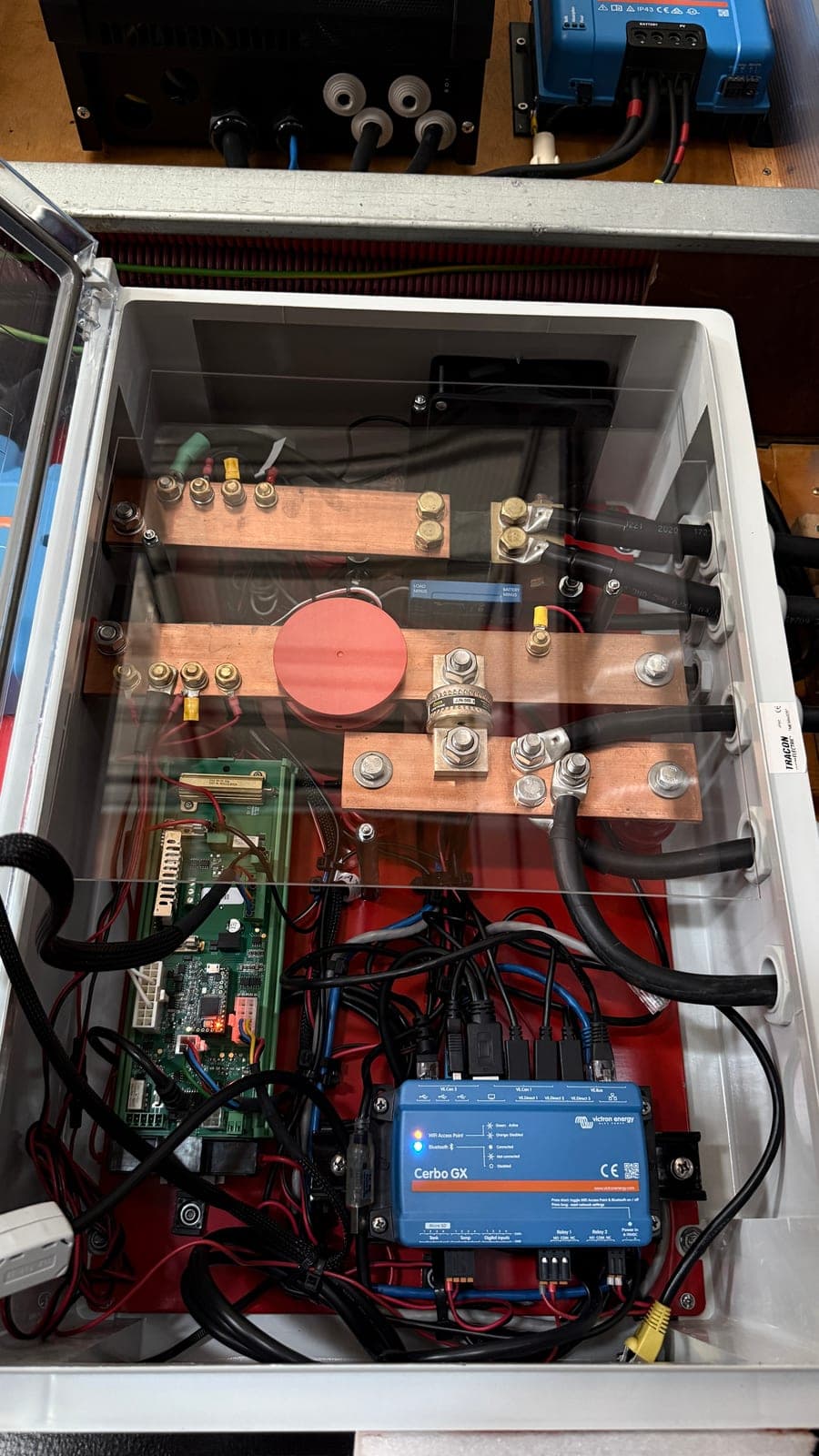 Caixa de distribuição de corrente contínua com tampa transparente levantada, mostrando três barramentos de cobre, um fusível cilíndrico de grande calibre, um shunt de medição e uma placa eletrónica de gestão de bateria.