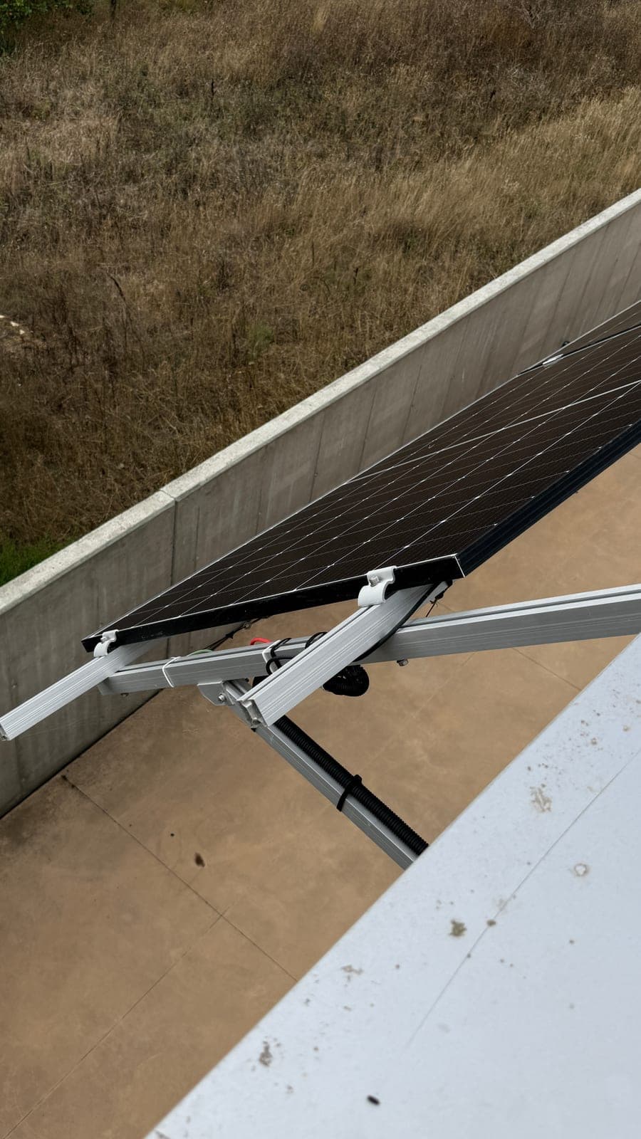 Bordo de um painel solar preso por um grampo branco a uma calha de alumínio, com a união aparafusada entre dois perfis e um tubo corrugado preto a correr por baixo, sobre um terraço de betão.