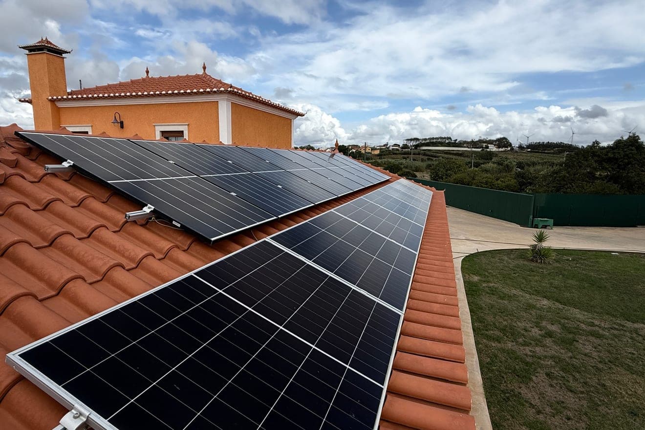 Duas fiadas de painéis solares assentes sobre um telhado de telha cerâmica cor de barro, a acompanhar a inclinação da cobertura, com uma chaminé, relva e um céu de nuvens ao fundo.