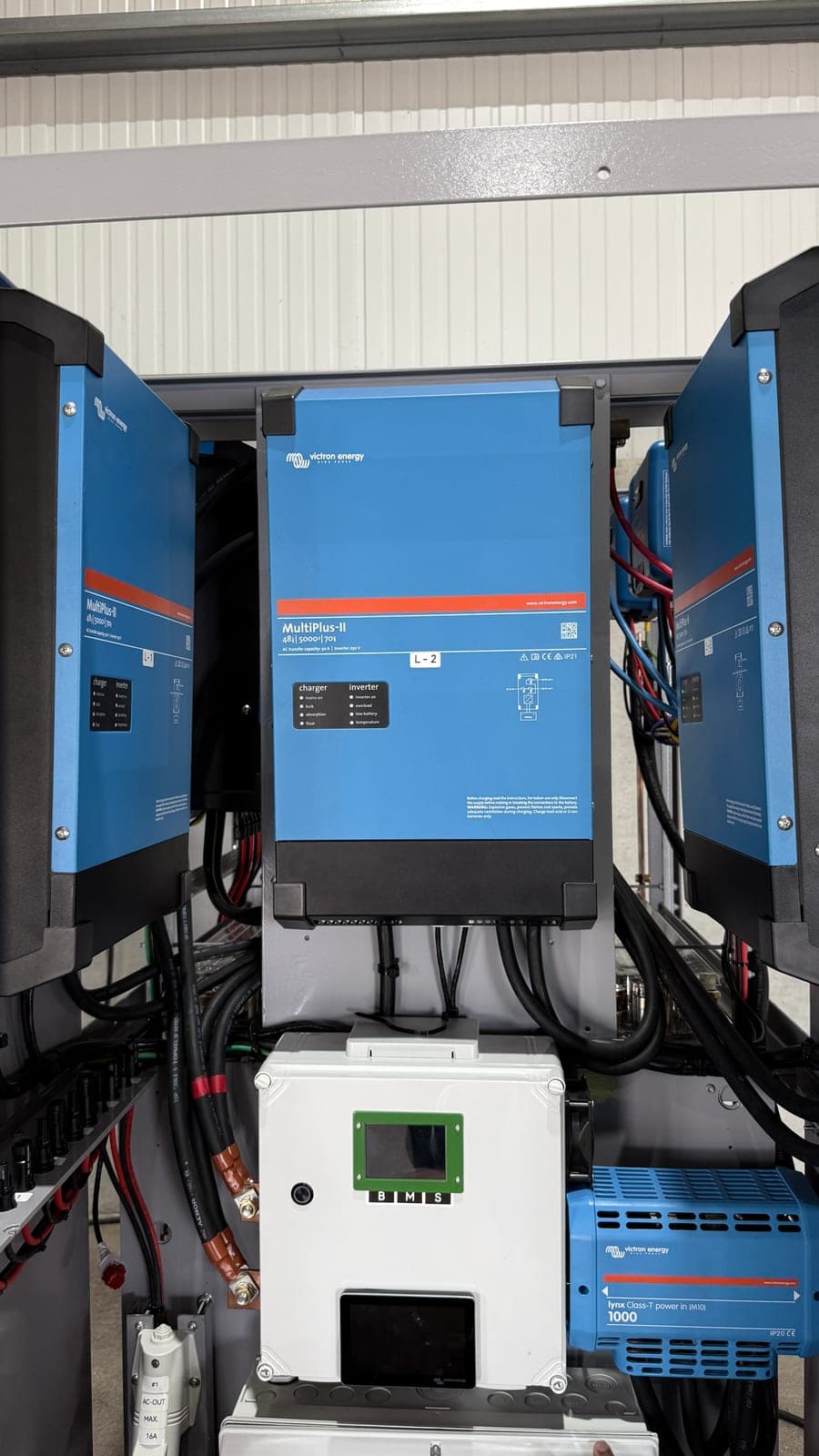 Três inversores Victron MultiPlus-II montados lado a lado e etiquetados L-1, L-2 e L-3, com uma caixa de gestão de bateria, um ecrã de leitura e um barramento de entrada por baixo.