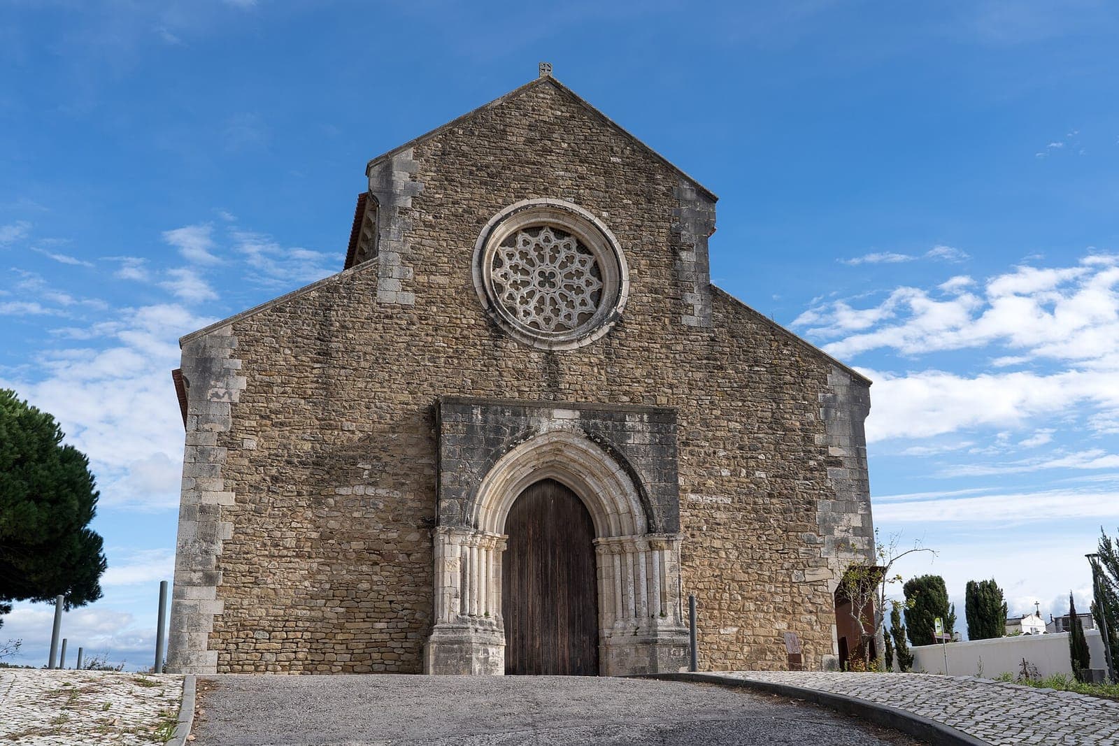 Fachada de pedra da Igreja de Santa Maria do Castelo, na Lourinhã, com portal gótico em arco e uma rosácea por cima, sob um céu azul com nuvens.