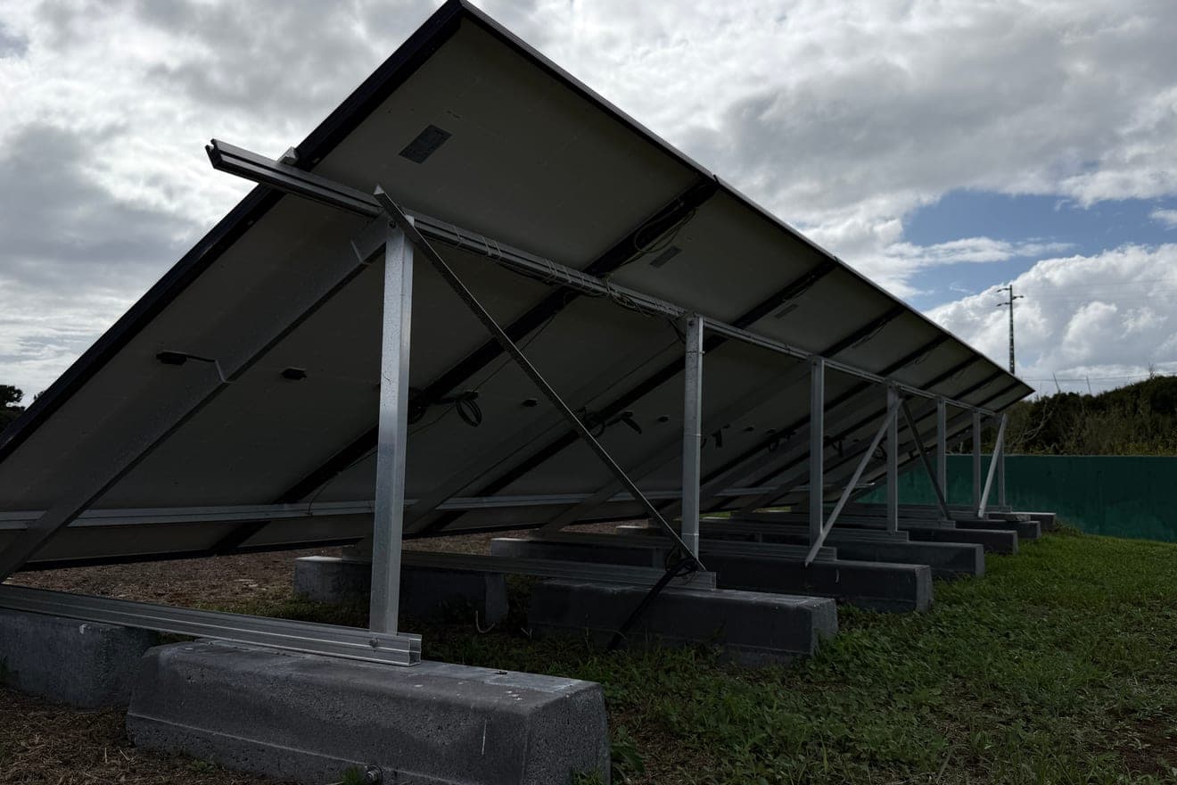 Estrutura metálica triangular vista por trás, com escoras na diagonal assentes em maciços de betão sobre relva, a suportar uma fila de painéis solares inclinados contra um céu de nuvens.