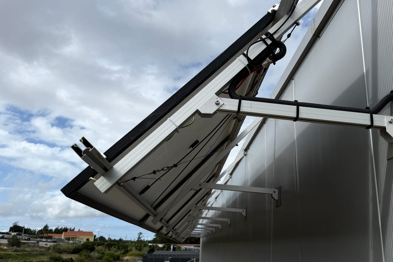 Painéis solares a sair na horizontal de uma parede vertical de chapa metálica, vistos por baixo, presos por apoios em esquadria aparafusados à parede, com uma calha de alumínio e um tubo corrugado a correr por baixo, contra um céu de nuvens.