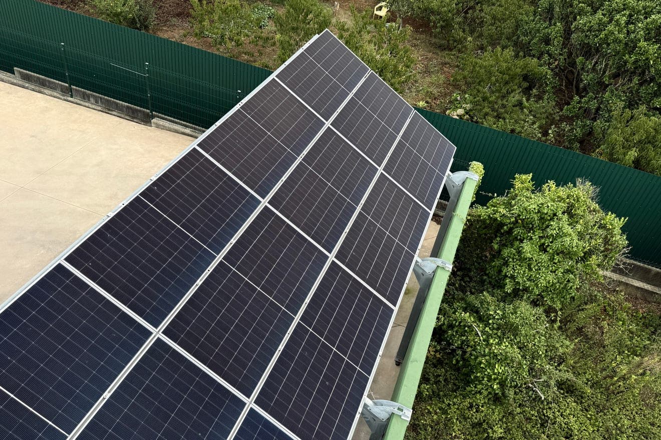 Fiada de painéis solares inclinada sobre um terraço de betão claro, presa por grampos a uma viga metálica pintada de verde, com uma vedação verde e vegetação densa do lado direito.