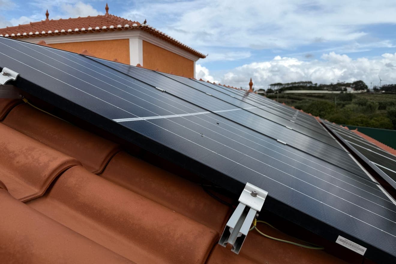 Painéis solares assentes sobre um telhado de telha cerâmica cor de barro, vistos de baixo e quase ao nível da cobertura, com um apoio metálico a assentar numa telha e a fachada da casa ao fundo.